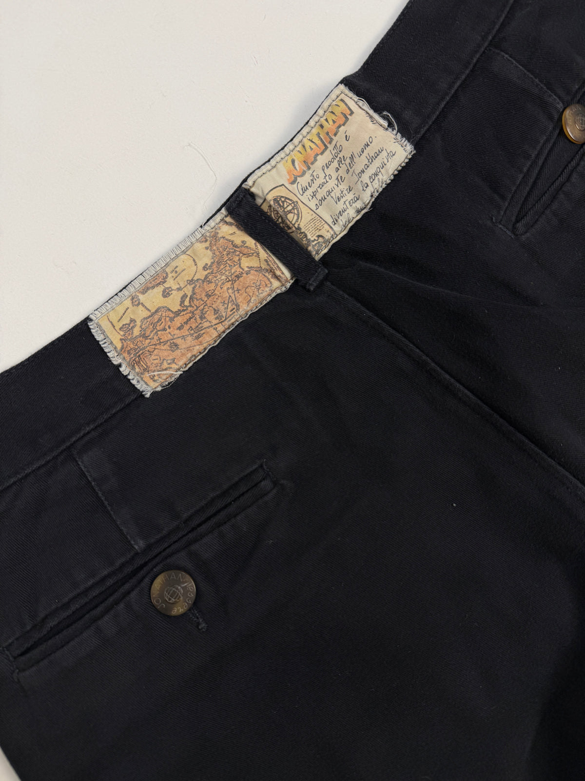 Dettaglio macro zip e pinces - zip metallo dorato con guardia nera, bottone singolo ottone logo ancora, doppia pinces profonda anteriore su cotone twill nero