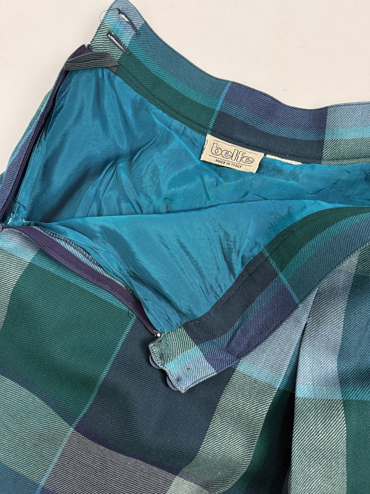 Dettaglio macro vita frontale - tasca a filetto verticale destra, 2 bottoni circolari resina blu navy fascia vita, tessuto twill tartan verde teal armatura diagonale visibile