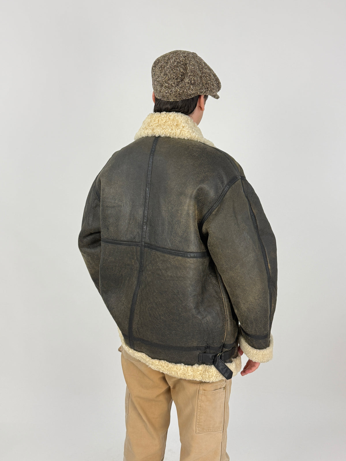 Dettaglio macro B-3 bomber - nappa montone pebbled grana pronunciata patina marrone ebano-oliva, patta tasca bassa rettangolare cucitura vista, fascetta laterale pelle scura fibbia bronzo D-ring, polsino shearling riccio avorio